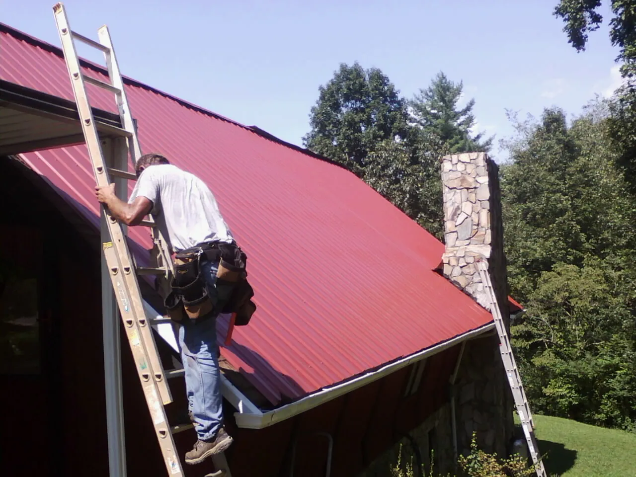 Roofer on a ladder beside a red metal roof and stone chimney — Soffit & Fascia Repair by Roofing Brothers in Argyle